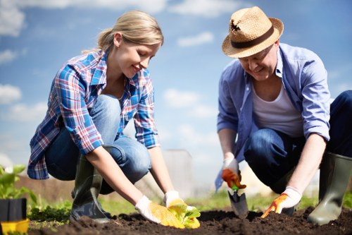 A young woman and an older woman are working together in a garden, crouched close to the soil, planting or tending to a small flowering plant. The young woman has blonde hair and is dressed in a blue plaid shirt and jeans, while the older woman wears a sun hat, a light blue shirt, and dark trousers. They are both wearing gardening gloves and appear to be engaged in outdoor gardening activities, with their focus on the plant and soil. The garden setting features dark, well-tilled soil, bordered by a variety of plants, with a backdrop of a clear blue sky and some distant trees or bushes. This scene demonstrates outdoor gardening and lawn care, emphasizing careful planting and horticultural work typical of professional gardening services in Uxbridge or surrounding areas served by Lawn Mowing Uxbridge. The natural lighting suggests a bright, sunny day, ideal for gardening or landscaping projects.