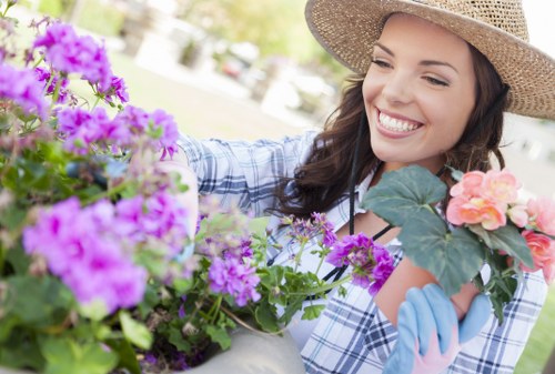 A woman wearing a straw sun hat and gardening gloves is smiling while tending to a variety of flowering plants in a well-maintained garden. The garden features lush green grass, bordered by flower beds filled with vibrant pink, purple, and peach blossoms, along with green foliage. In the background, there are mature trees, neatly trimmed hedges, and a paved patio area, suggesting a landscaped outdoor space in a residential garden near Uxbridge. The scene captures a bright, sunny day with natural daylight illuminating the colorful flowers and greenery, reflecting a healthy outdoor environment suitable for gardening and lawn care services provided by Lawn Mowing Uxbridge.