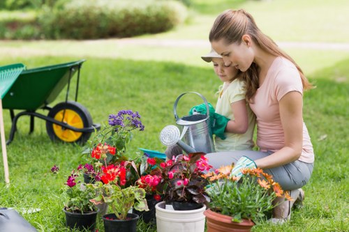 A young woman and a small child are kneeling together on a well-maintained, lush green lawn in a garden, engaging in gardening activities. The foreground features numerous potted plants and flower beds with vibrant red, pink, purple, and orange blossoms, along with green foliage, all arranged around them. The woman is wearing a light pink top and blue jeans, and she is holding a watering can, with her attention directed towards the plants. The child, dressed in a yellow shirt and green gardening gloves, is standing close by, observing the plants with interest. In the background, a green yard cart with yellow wheels is visible, along with a soft-focus hedge and trees that border the backyard, indicating a landscaped outdoor space typical of Uxbridge gardens. The weather appears bright and clear, with sunlight illuminating the scene and casting gentle shadows, creating a welcoming outdoor environment suitable for gardening and lawn care services offered by Lawn Mowing Uxbridge, which manages local gardening needs in the area around UB8 postal code.